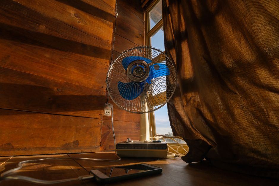 A blue-bladed electric fan hanging by a window against a wooden plank wall